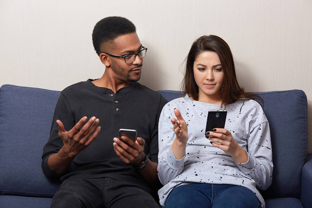 Studio shot of couple sitting together on comfortable sofa in apartment and using mobile phone, looking on screen, dark skinned male and caucasian female wearing casually, reading something unpleasant - Foto, immagini