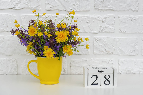Wooden cubes calendar July 28 and yellow cup with bright colored flowers against white brick wall. Template calendar date for your design Copy space - Foto, imagen