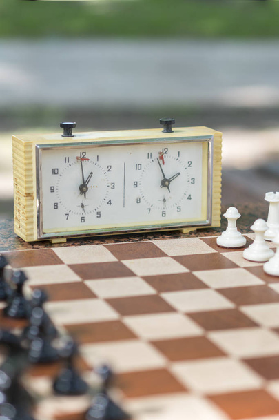 Szachownica z kawałkami i zegar na drewnianym biurku w związku z turnieju szachowego. Turniej Szachowy z zegarem szachowym na drewnianym stole. Zdjęcie pionowe - Zdjęcie, obraz