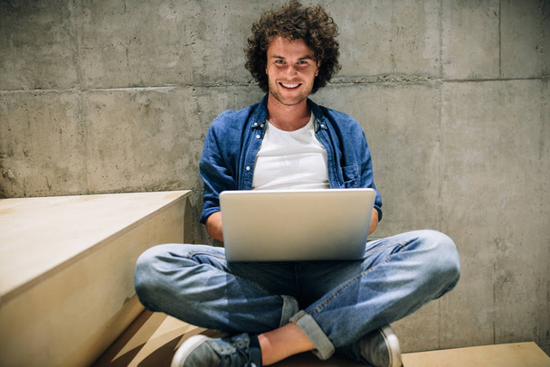 Smart young man with curly hair using laptop for working online, surfing on Internet, sitting on the stairs at concrete wall. Handsome male reading and typing on his laptop computer in the office - Fotografie, Obrázek
