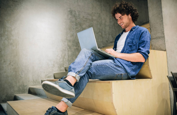 Indoor shot of young man with curly hair using laptop for chatting online with friends, browsing Internet, sitting on the stairs at concrete wall. Candid male reading and typing on his laptop computer - Valokuva, kuva