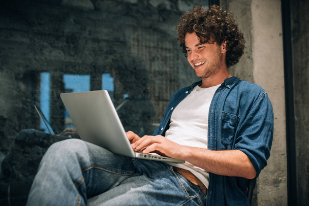 Portrait of happy young man with curly hair using laptop for working online. Smart freelance man chatting online with clients. Handsome male reading news, typing on his laptop computer in the office - Fotoğraf, Görsel