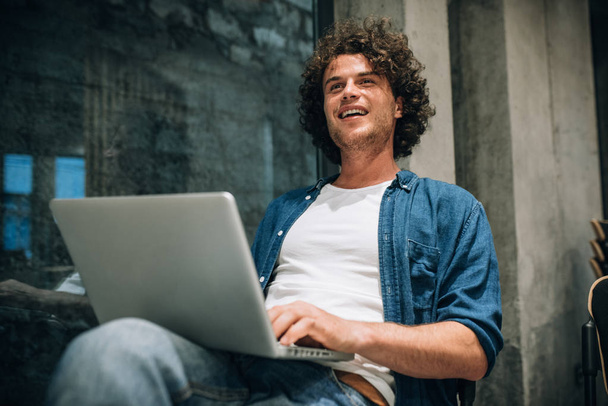 Handsome young man with curly hair using laptop for working online. Smart freelance man chatting online with clients. Smart male reading news, typing on his laptop computer in the modern office - Фото, зображення