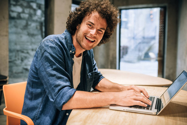 Portrait of happy young man with curly hair using laptop for working and browsing online. Smart Caucasian male reading news, typing on his laptop computer in the modern office. Business and people - Fotó, kép