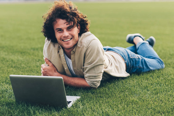 Handsome young freelance businessman sitting on the ground, working on laptop. Smiling man with curly hair using laptop for chatting online, sitting on the grass in the park. People and business - Photo, Image