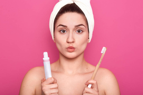 Studio shot of pleased female model pouting lips, wears white towel on head, ready for brushing teeth with toothbrush and toothpaste, cares of hygiene and beauty, isolated over pink background. - Фото, изображение