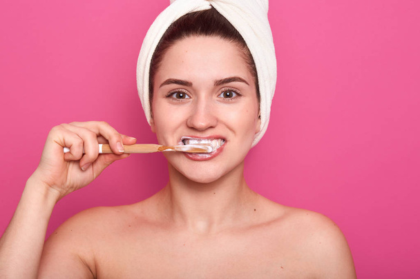 Woman with tooth brush, cleaning her teeth, posing in bathroom with bare head and white towel, female doing morning procedures after taking shower, isolated on rosy background. Hygiene concept. - Photo, image