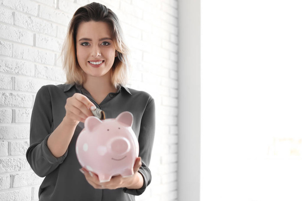 Beautiful woman putting money into piggy bank near brick wall. Space for text - Zdjęcie, obraz