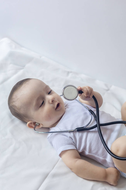 Küçük sevimli bebek doktoru. 6 aylık çocuk stetoskop ile oynuyor. Çocuk bir doktor gibi eğleniyor - Fotoğraf, Görsel