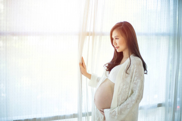 Νέοι Ασίας όμορφη έγκυος γυναίκα στέκεται κοντά στο παράθυρο - Φωτογραφία, εικόνα