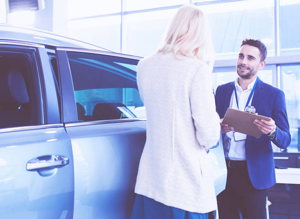 Beautiful young woman is talking to handsome car dealership worker while choosing a car in dealership - Φωτογραφία, εικόνα