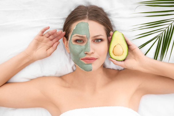 Beautiful woman holding avocado near her face with clay mask on white fabric, above view - Photo, image