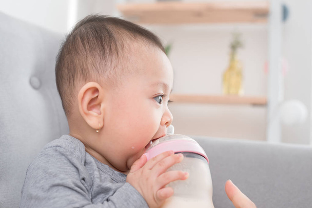 Schattige babymeisje zit in kamer op Bank drinken melk uit de fles en lachend. Gelukkig kind. Familie mensen binnen interieur concepten. Beste tijd voor jeugd! - Foto, afbeelding