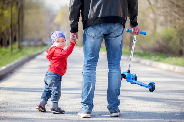 Father and toddler son going for a walk holding hands, view from behind. Dad is carrying little blue kick scooter, son half turned back and is looking back. Family leisure outdoors concept - Zdjęcie, obraz