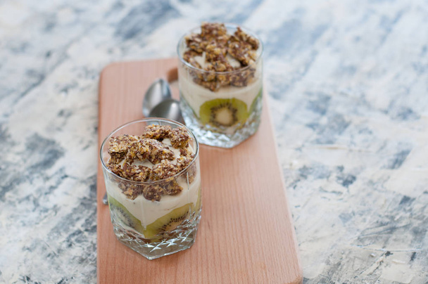 Granola, kiwi, banana and Greek yogurt in two glass cups on wooden board gray concrete, with spoons, copy space, view from the top and from the side. Fitness, figure, body and healthy food - Foto, Imagen