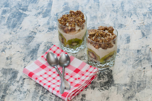 Deser Granola, grecki jogurt, kiwi i banan w dwóch szklanek kubki z łyżkami i różowy ręcznik w kratkę na szarym betonie. Fitness, figura, ciało i zdrowa żywność - Zdjęcie, obraz