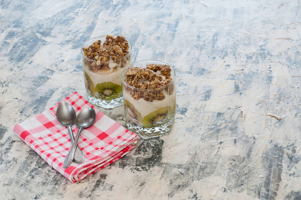 Deser Granola, grecki jogurt, kiwi i banan w dwóch szklanek kubki z łyżkami i różowy ręcznik w kratkę na szarym betonie. Fitness, figura, ciało i zdrowa żywność - Zdjęcie, obraz