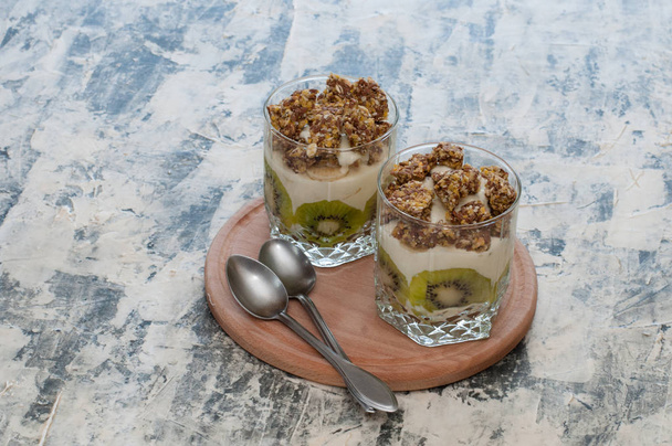 Deser Granola, grecki jogurt, kiwi i banan w dwóch szklanek kubki z łyżkami na drewnianej okrągłej płycie, szary betonu. Fitness, figura, ciało i zdrowa żywność - Zdjęcie, obraz