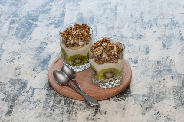 Deser Granola, grecki jogurt, kiwi i banan w dwóch szklanek kubki z łyżkami na drewnianej okrągłej płycie, szary betonu. Fitness, figura, ciało i zdrowa żywność - Zdjęcie, obraz