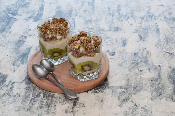 Dessert granola, Greek yogurt, kiwi and banana in two glass cups with spoons on wooden round board, gray concrete. Fitness, figure, body and healthy food - Foto, Imagem