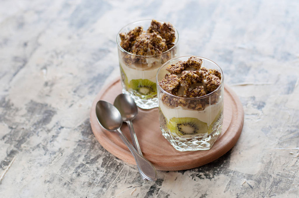 Dessert granola, Greek yogurt, kiwi and banana in two glass cups with spoons on wooden round board, gray concrete, close-up. Fitness, figure, body and healthy food - Valokuva, kuva