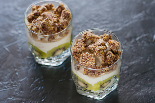 Greek yogurt, granola, banana and kiwi. Two desserts in glass cups on dark background. Concept body and healthy food - Fotoğraf, Görsel