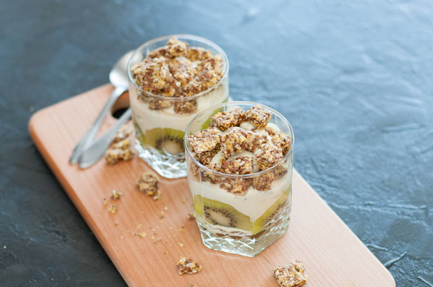 Greek yogurt, granola, banana and kiwi. Desserts in glass cups with spoons on wooden platform, dark background, view from the top. Concept body and healthy food - Photo, Image