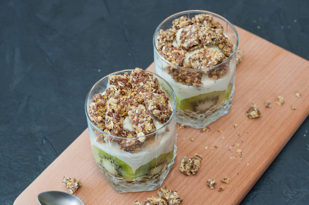 Greek yogurt, granola, banana and kiwi. Desserts in glass cups with spoons on wooden platform, dark background, view from the top. Concept body and healthy food - Foto, Bild