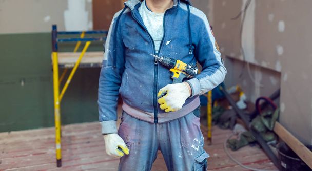 Worker with protective gloves is holdind a perforator with some drywall screws in apartment that is under construction, remodeling, renovation, overhaul, extension, restoration and reconstruction - Zdjęcie, obraz