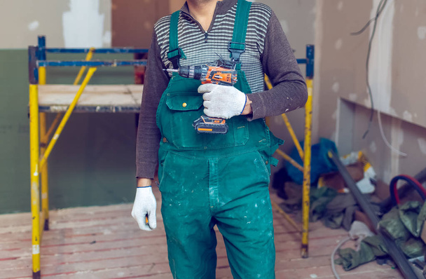 Worker with protective gloves is holdind a perforator with some drywall screws in apartment that is under construction, remodeling, renovation, overhaul, extension, restoration and reconstruction - Photo, image