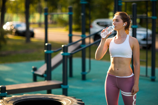 Jeune femme caucasienne s'entraîne sur l'aire de jeux extérieure et l'eau potable de la bouteille. Crossfit et entraînement de fitness à l'extérieur
. - Photo, image