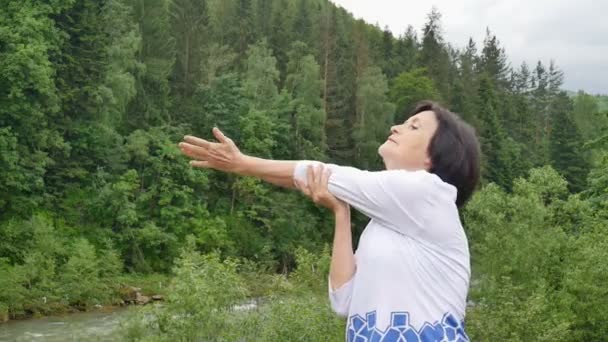 Mujer mayor con el pelo corto y oscuro haciendo un ejercicio de estiramiento para relajarse en la mañana fuera sobre el paisaje de bosque y montañas
 - Imágenes, Vídeo