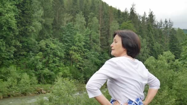 Senior vrouw met korte donkere haren doen een stretching oefening voor ontspanning in de ochtend buiten over landschap van bos en bergen - Video