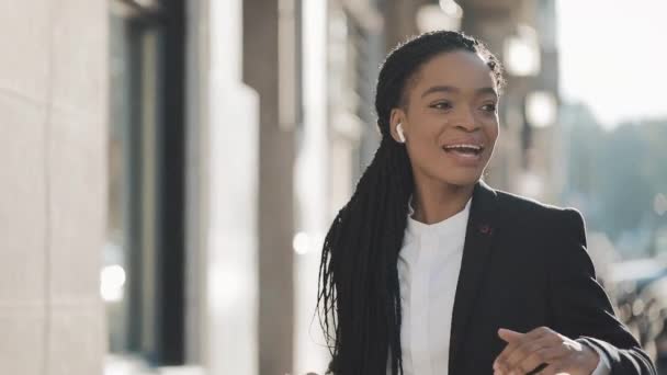 Young happy African American businesswoman in earphones walking the city street while listening to the music on the smartphone and dancing. Outdoor. - Filmagem, Vídeo