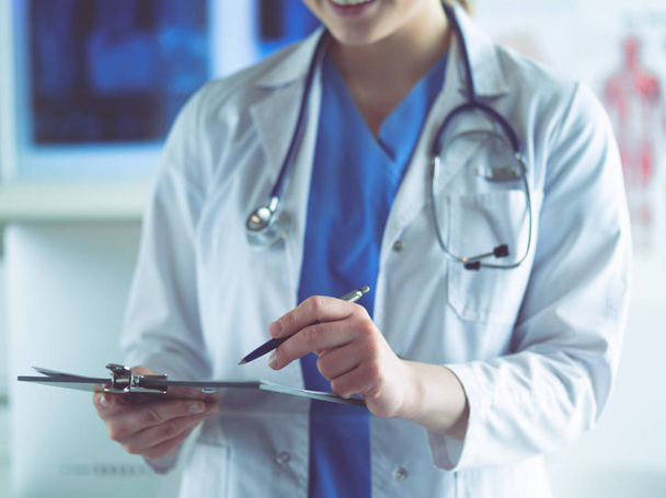 Dokter met een stethoscoop, met een notitieboekje in zijn hand. Close-up van een vrouwelijke arts die een medisch formulier invult aan het klembord terwijl hij recht in het ziekenhuis staat - Foto, afbeelding