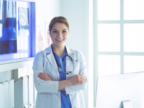 Νεαρή γυναίκα γιατρός με λευκή στολή στέκεται στο γραφείο κλινικές - Φωτογραφία, εικόνα