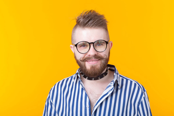 Joven hombre de moda positivo hipster con una barba bigote y collar fetiche en camisa posando sobre un fondo amarillo. Concepto de roca y subcultura
. - Foto, Imagen