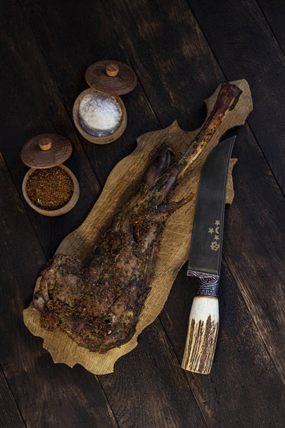 Baked goat leg on oak table with spices and knife - Фото, зображення