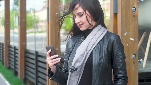 привлекательная молодая девушка в стильном сером шарфе и черном Курске смотрит на телефон и нажимает на кнопки пальцами. Длинные волосы развиваются при сильном ветре. Городские здания на заднем плане
 - Кадры, видео
