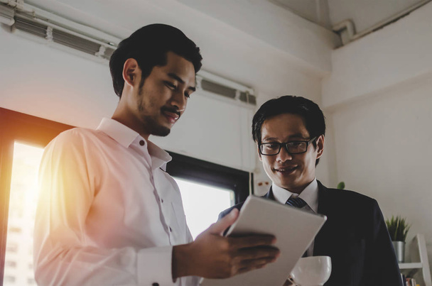 two asian young partner business man investor talking and reading information about financial statistics in mobile tablet together standing in office, finance, investment, digital technology concept - 写真・画像