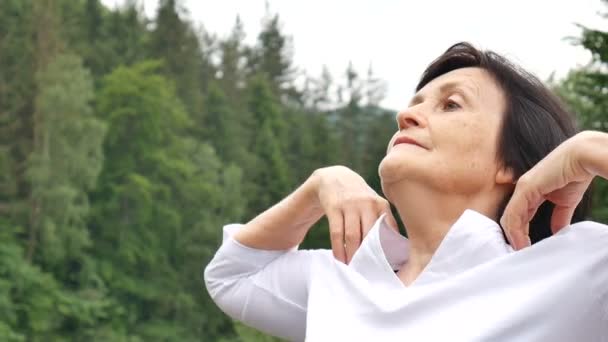 Senior vrouw met korte donkere haren doen een stretching oefening voor ontspanning in de ochtend buiten over landschap van bos en bergen - Video