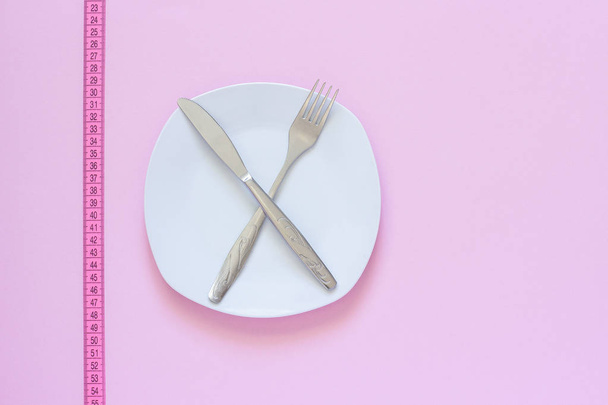Crossed fork and knife on white plate and tape measure on pink background. - Φωτογραφία, εικόνα