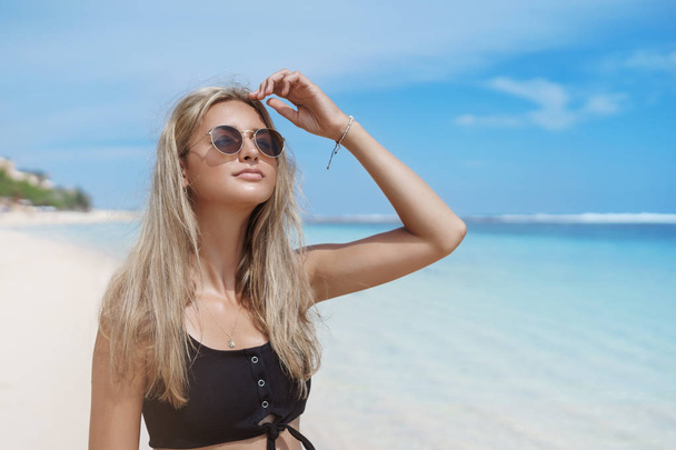 Girl gets perfect tan during summer vacation Bali. Attractive relaxed carefree blond woman wear sunglasses cropped top close eyes raise head sun, walk sandy beach contemplating ocean, surfers - Fotografie, Obrázek