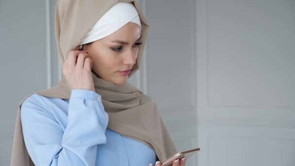 Мусульманка, носящая хиджаб, слушает музыку в смартфоне с помощью беспроводного наушника
. - Кадры, видео