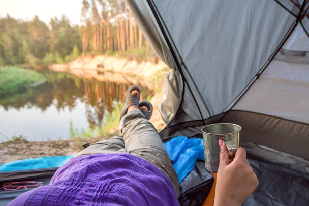 Feet woman relaxing enjoying coffee. River view from tent camping entrance. Travel concept adventure vacations outdoor - Foto, afbeelding
