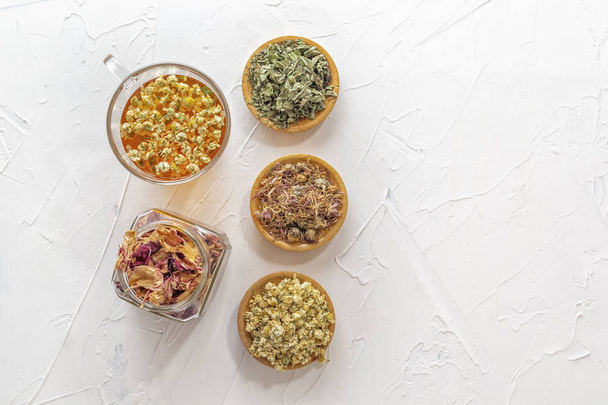 Chá de ervas com camomila farmacêutica, crisântemo seco, um frasco de pétalas de rosa seca e caçadores em uma mesa branca
 - Foto, Imagem