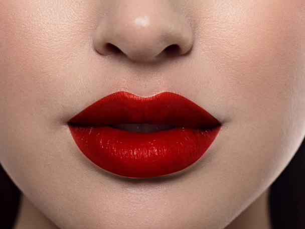 Cosmetics, makeup and trends. Bright lip gloss and lipstick on lips. Closeup of beautiful female mouth with red lip makeup. Beautiful part of female face. Perfect clean skin in red light - Zdjęcie, obraz