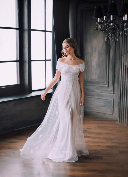 gentle princess in long light white dress with open shoulders in dark black room in Gothic style, fabulous girl, vintage gown, hall with large windows, beauty sharpened in mysterious castle - Photo, Image