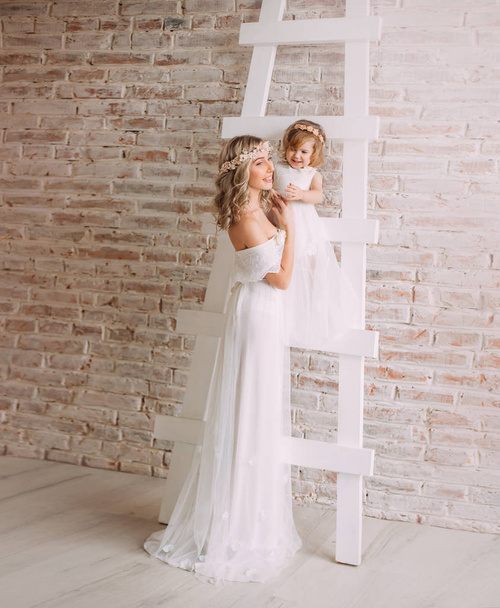 joyful happy woman in long white lace dress with floral wreath on her head, mother holding her beloved little daughter in loose gown on light stairs near brick wall. positive emotions with children - Photo, Image