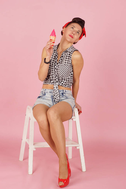 Young beautiful brunette sits and dreams with a cone of ice cream in her hand, pin-up style on a pink background - Foto, afbeelding